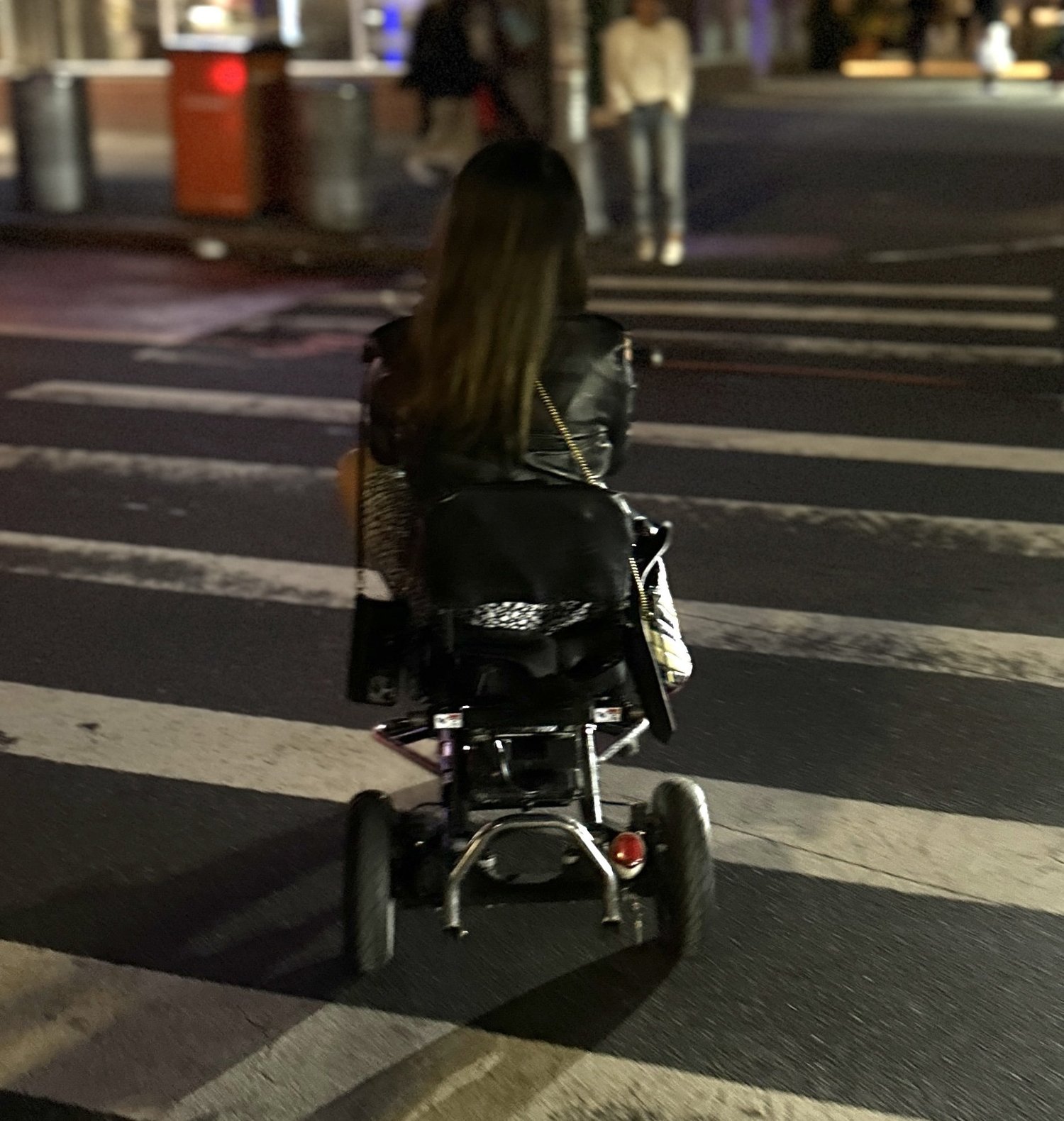 Back image of Sarah Kim crossing a city street at a crosswalk in her mobility scooter at night.